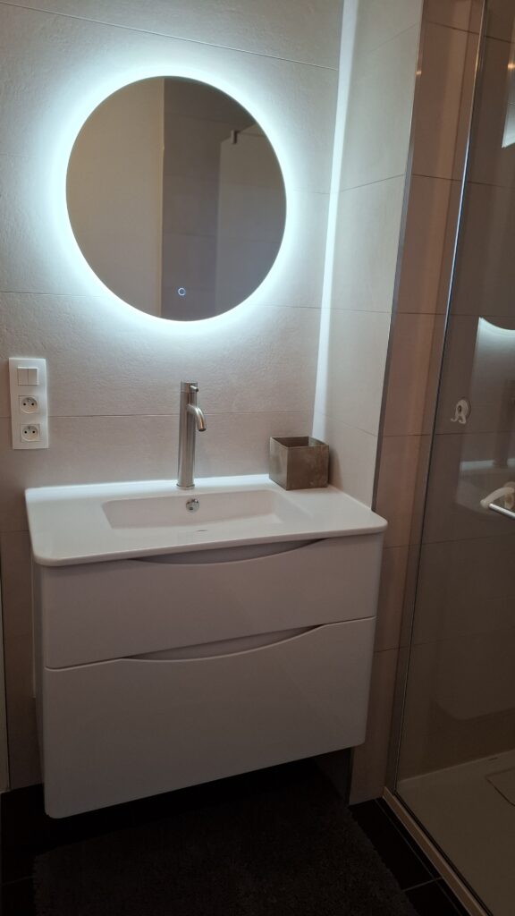 Meuble vasque suspendu blanc avec tiroirs, robinetterie chromée et miroir rond rétroéclairé LED dans une salle de bain moderne et épurée.