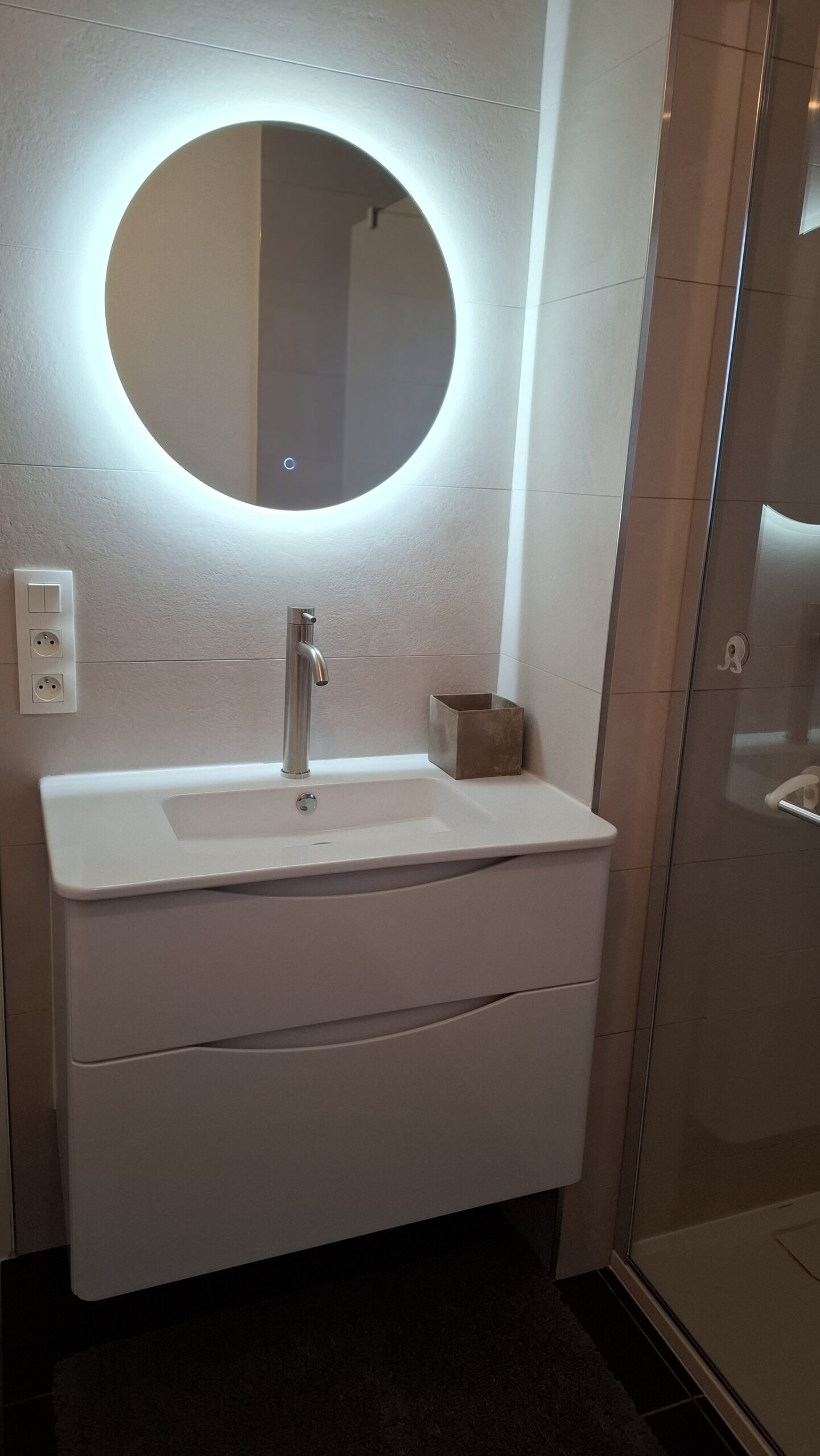 Meuble vasque suspendu blanc avec tiroirs, robinetterie chromée et miroir rond rétroéclairé LED dans une salle de bain moderne et épurée.